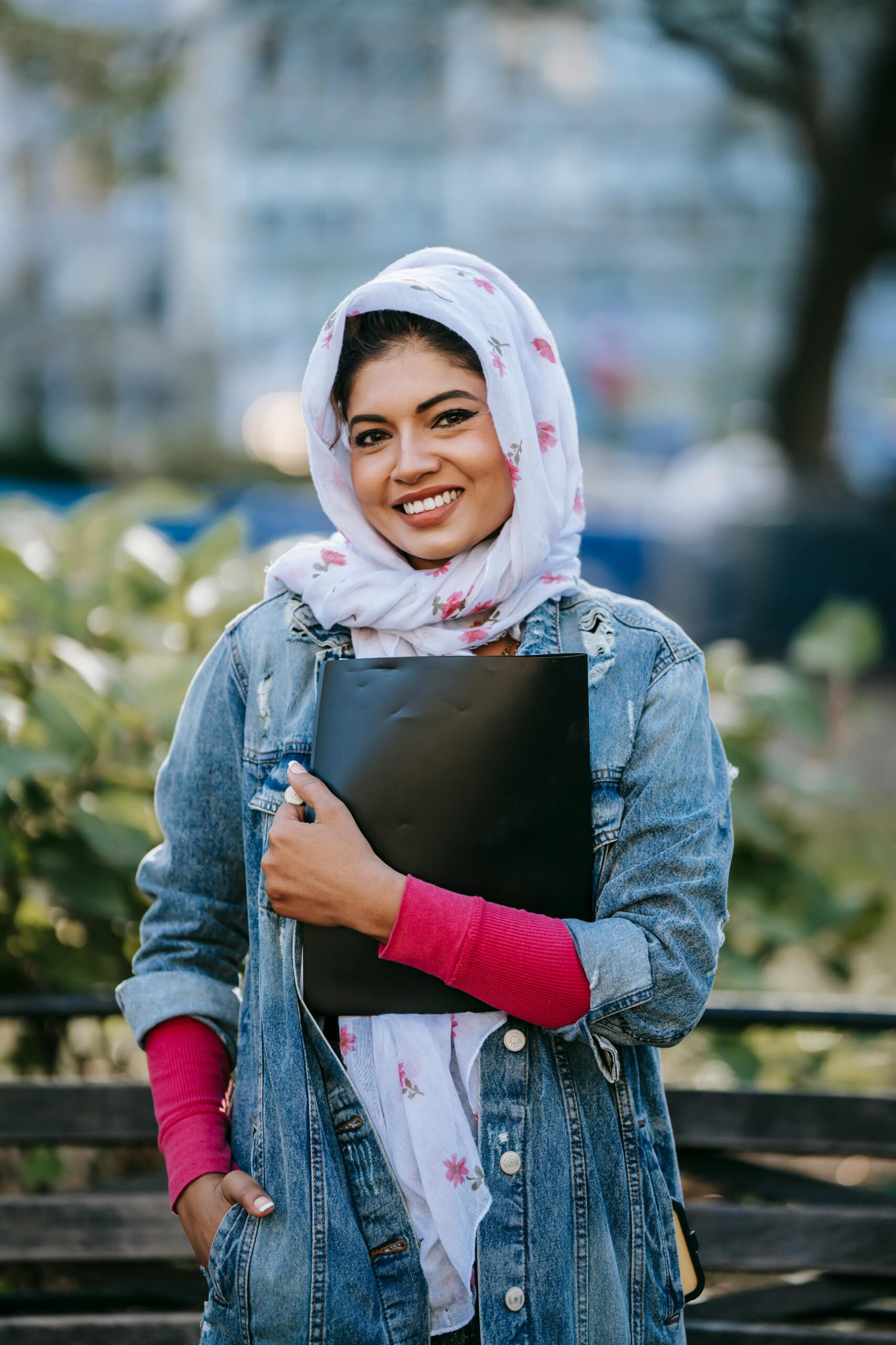 Positive young Muslim woman in hijab and jeans coat standing with black folder near wooden railing of city park on sunny day and looking at camera